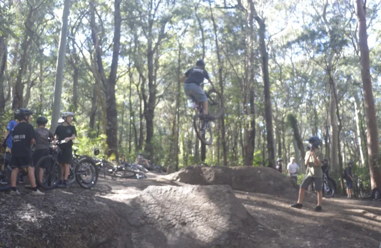 Mountain-bike rider jumping a dirt feature while riders watch from the forest trail