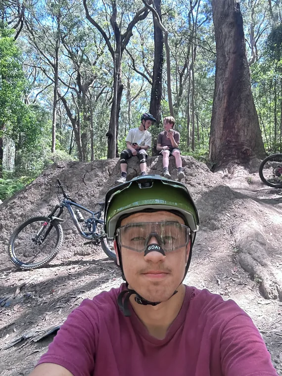 Helmeted rider taking a selfie on a wooded dirt trail, with two riders seated behind and mountain bikes nearby